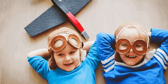 2 children lying down wearing pilot goggles with a toy plane
